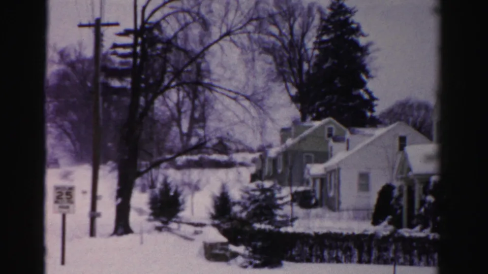 8mm home movie frame circa mid-1950s: snow-blanketed Albany, New York suburban street with modest one-story homes, bare trees, and a roadside sign on a utility pole.