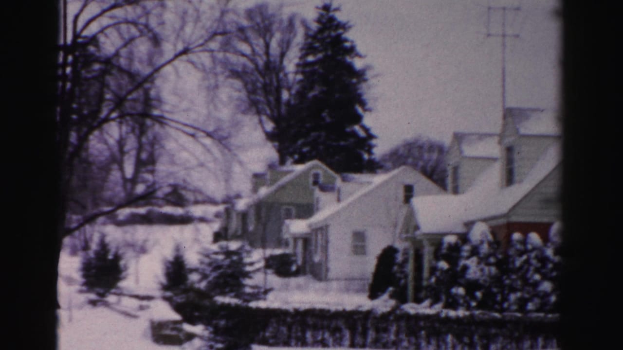 Snow-covered suburban homes with a prominent evergreen tree and utility poles; camera has panned slightly right from the opening frame.