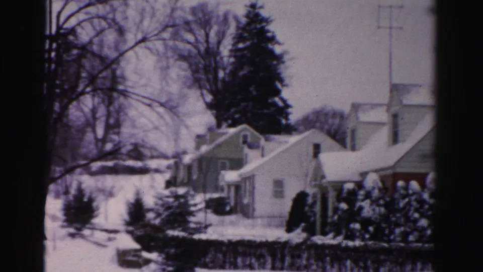 Mid-1950s 8mm home movie pan frame showing snow-covered one-story homes and an evergreen along a residential street in Albany, New York.