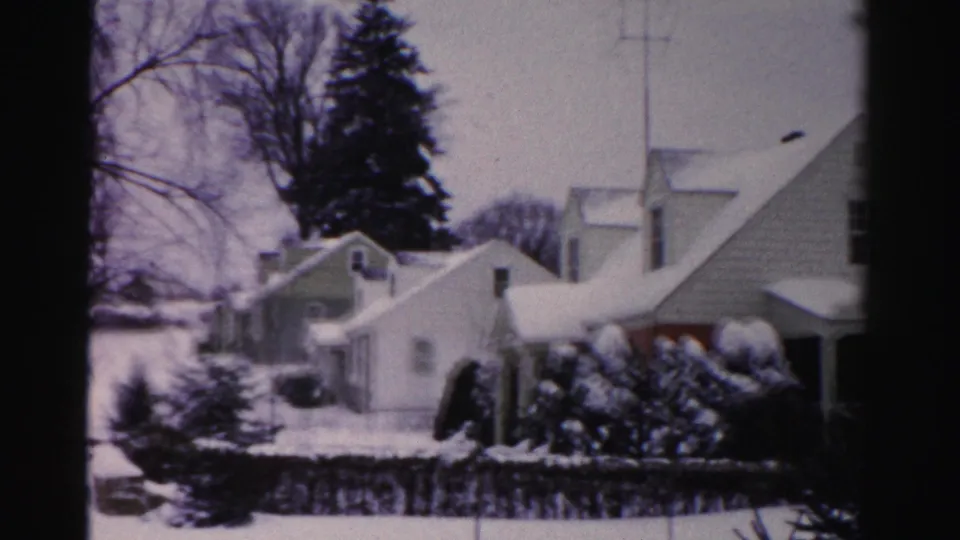 8mm home movie frame showing mid-1950s snow-covered suburban homes in Albany, New York, with an evergreen tree and a red-accented facade element visible.