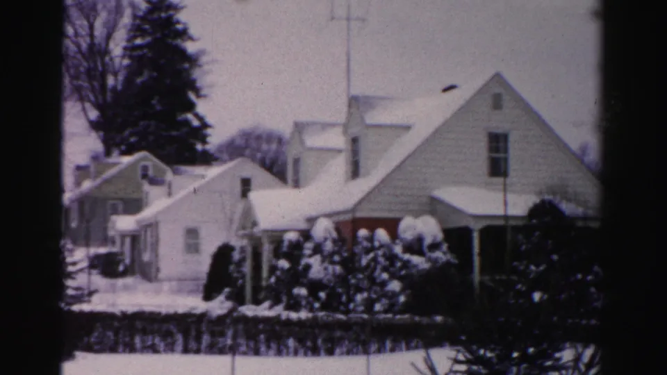 Final 8mm pan frame across a 1950s Albany, New York suburban street showing snow-covered gabled homes with snow-laden evergreen shrubs in the foreground.