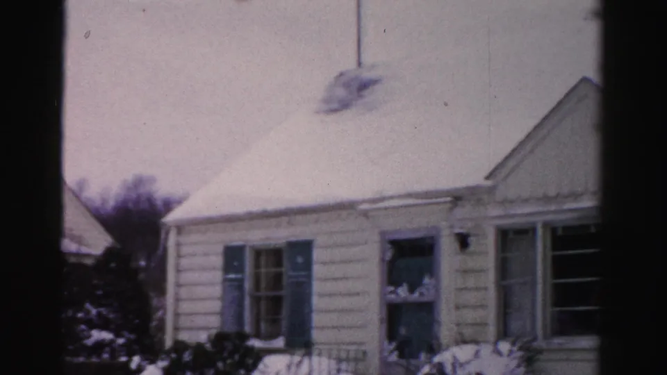 Pan across white clapboard house with blue-shuttered windows and snow-laden roof; Albany, New York area, circa mid-1950s, 8mm color film.