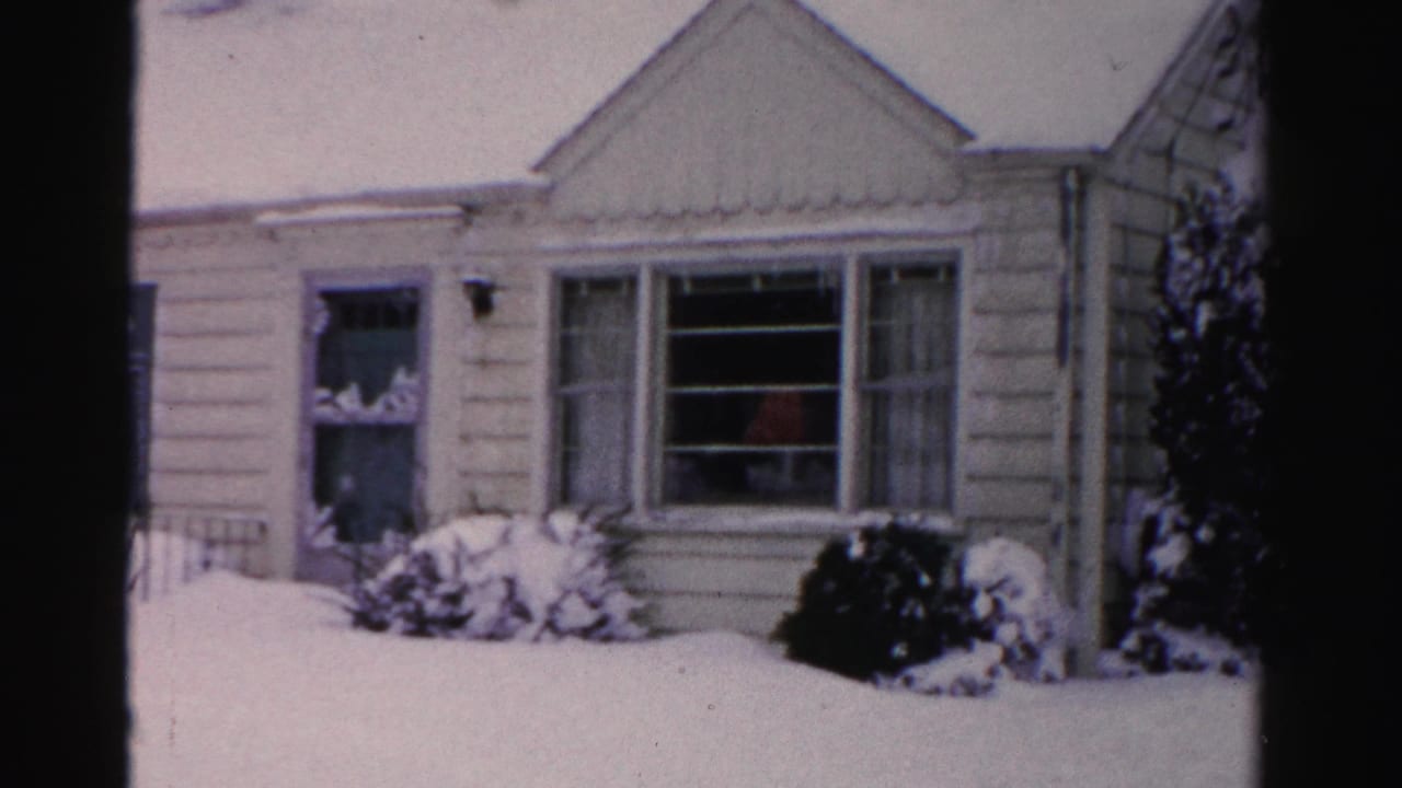 Center section of pale clapboard house with deep snow on roof and yard, faint reddish figure visible through window, mid-1950s Albany, New York, 8mm home movie.
