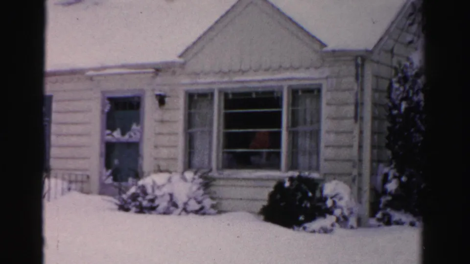Center section of pale clapboard house with deep snow on roof and yard, faint reddish figure visible through window, mid-1950s Albany, New York, 8mm home movie.