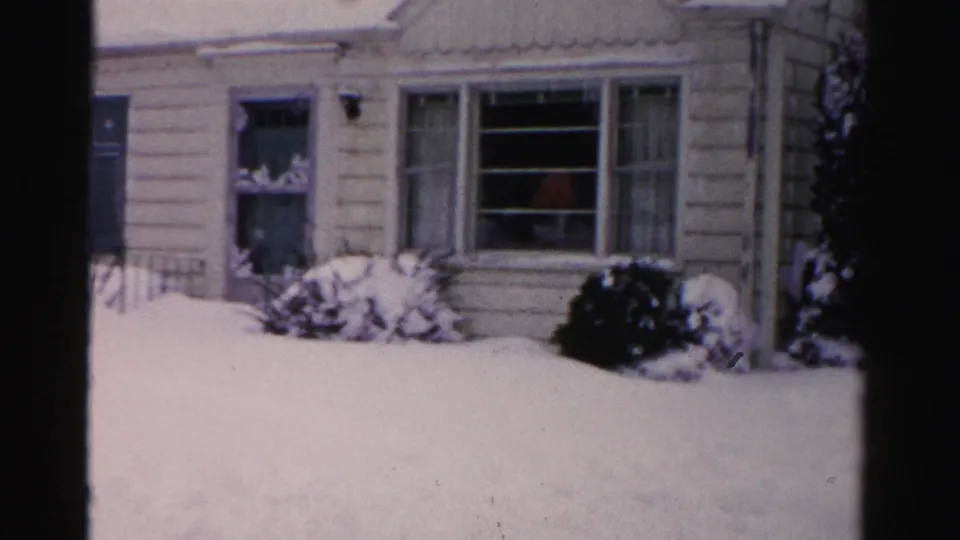 Frontal view of pale clapboard house with wall-mounted exterior lantern and deeply snow-laden shrubs, red figure faintly visible through windows, mid-1950s 8mm home movie.