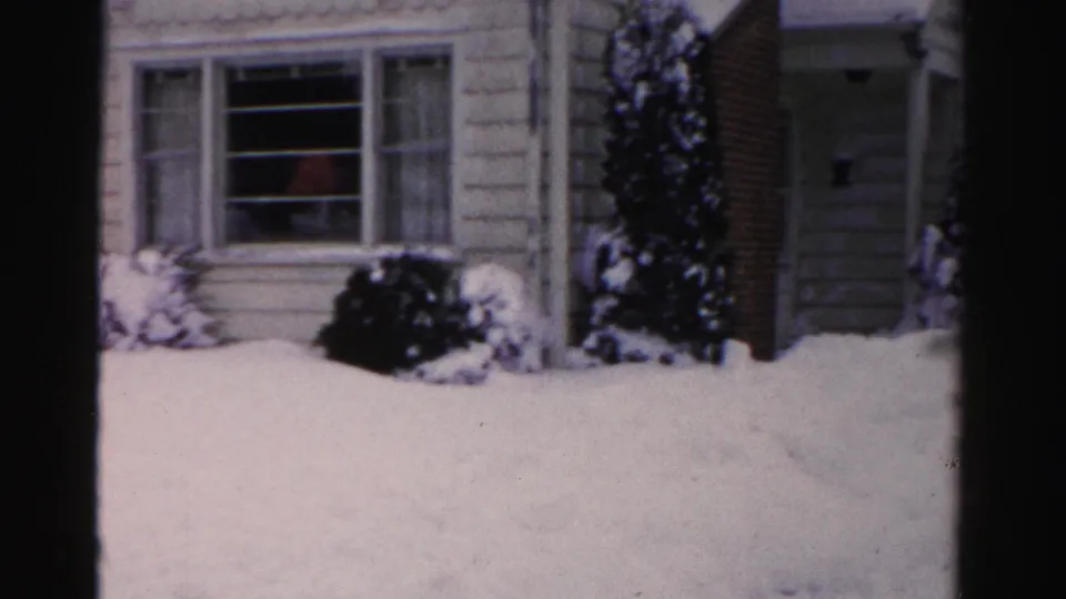 Right portion of pale clapboard suburban house with front entry area visible, snow-covered evergreen shrubs, faint red figure through window, mid-1950s Albany, New York, 8mm home movie.
