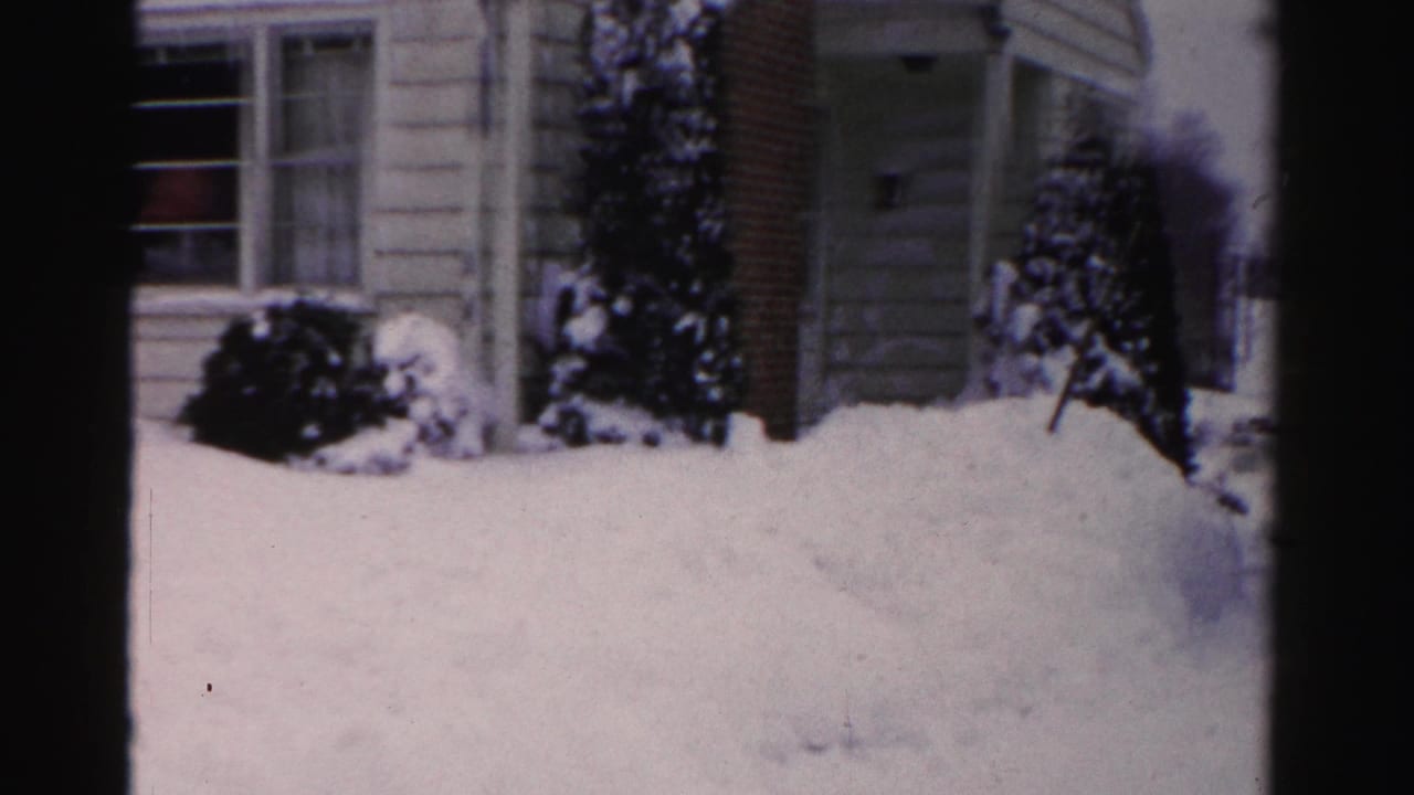 Snow-laden evergreen shrubs and a deep snow drift before a house section with horizontal siding, 8mm color film.