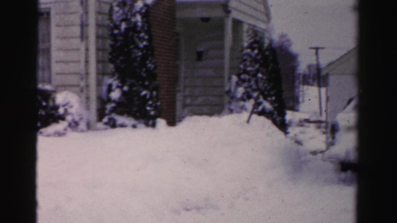 Snow-covered evergreen shrubs and deep drift beside a house exterior with a utility pole at right, Albany NY area, mid-1950s 8mm home movie.