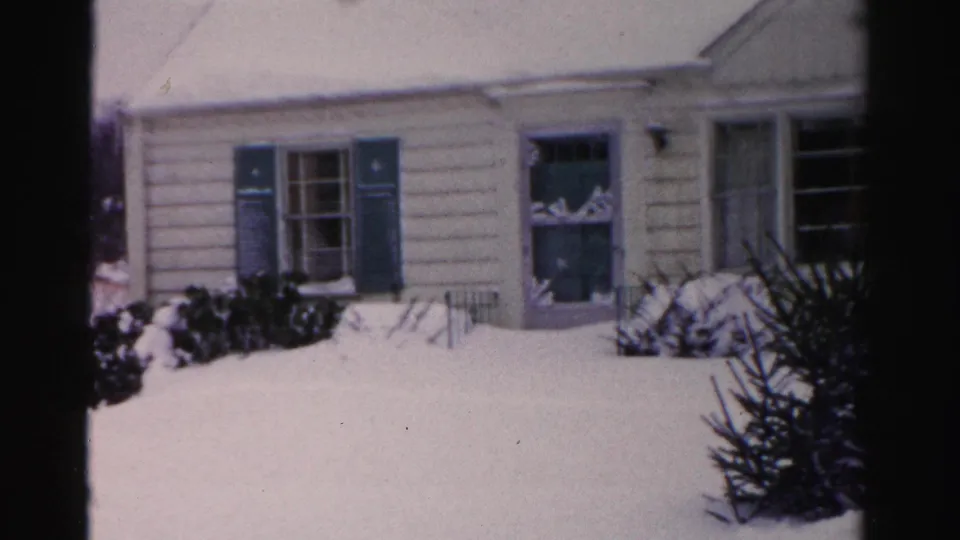 Ranch-style house with cream lap siding and blue-teal shutters under a snow-covered roof, Albany NY suburban setting, mid-1950s 8mm home movie.