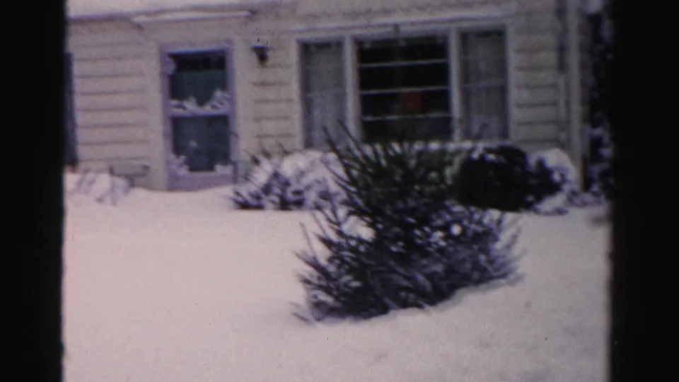 Ranch house facade with blue-teal shutters and cream lap siding; large snow-covered evergreen shrub in foreground, Albany NY area, mid-1950s 8mm home movie.