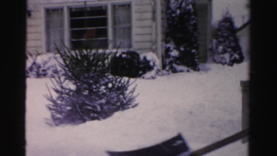 8mm home movie frame from a mid-1950s Albany, New York residential yard; snow-laden evergreen bushes and house facade, shovel blade in foreground.