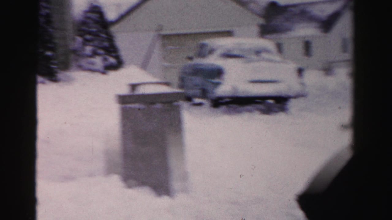 8mm frame showing a snow-covered driveway with what appears to be a mid-century automobile, metal trash cans, and neighboring houses, Albany, NY, mid-1950s.