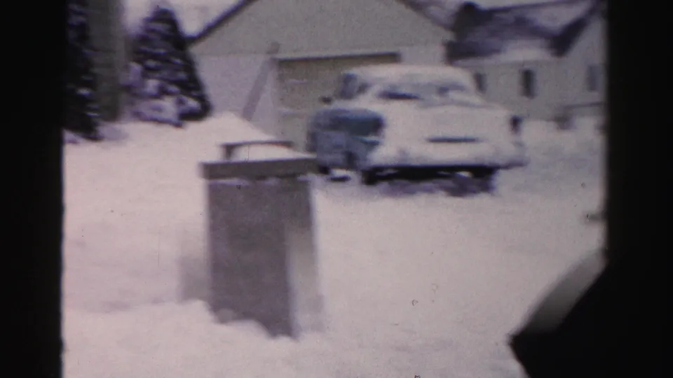 8mm frame showing a snow-covered driveway with what appears to be a mid-century automobile, metal trash cans, and neighboring houses, Albany, NY, mid-1950s.