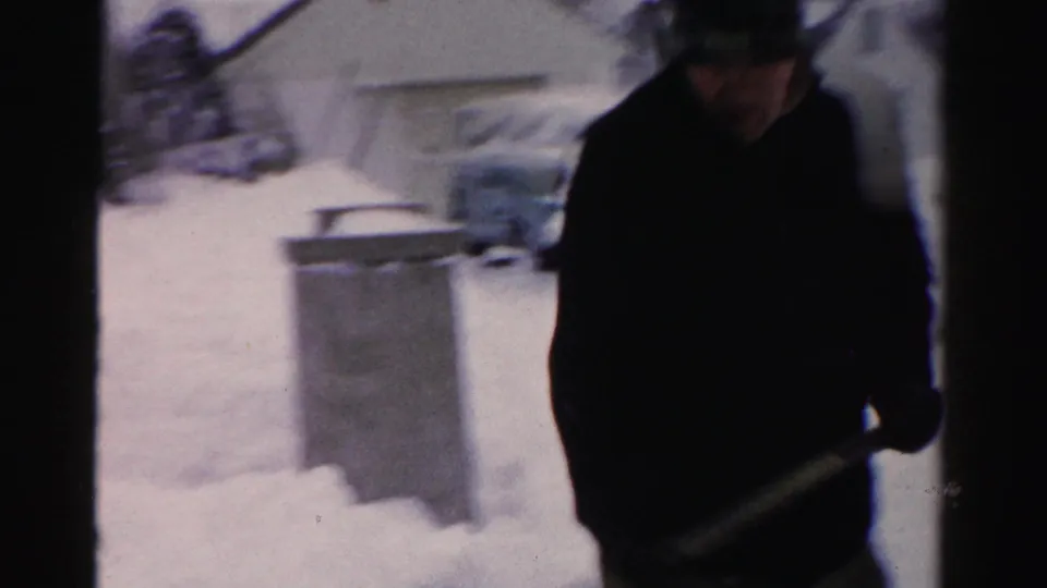 8mm home movie frame of a man in a dark coat and hat clearing snow from a driveway, mid-1950s Albany, New York; metal trash can and snow-covered surroundings in background.