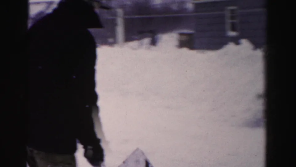 8mm close frame of a man in a dark winter coat with shovel, looking downward; fence posts and neighboring structure visible through snow, mid-1950s Albany, NY.
