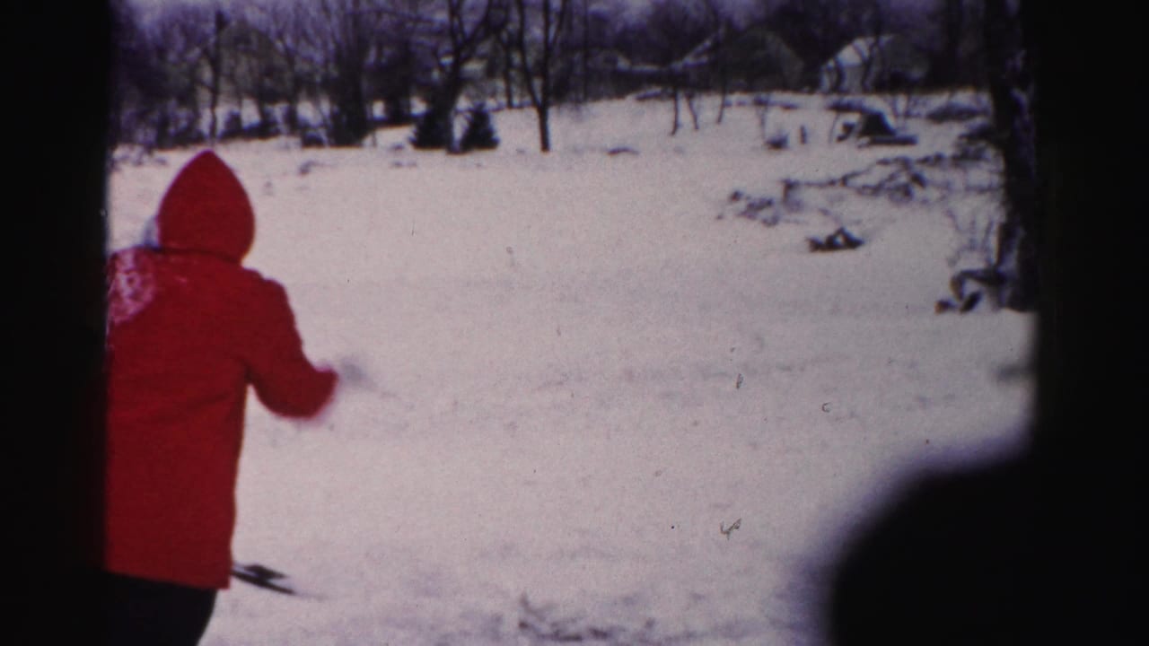 Adult in red hooded coat seen from behind in a snow-covered residential yard, consistent with mid-1950s Albany, New York 8mm home movie footage.