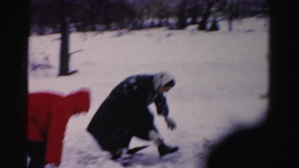 Adult in dark coat scooping snow in a residential yard with utility pole visible at rear, consistent with 1950s Northeast suburban 8mm home movie.