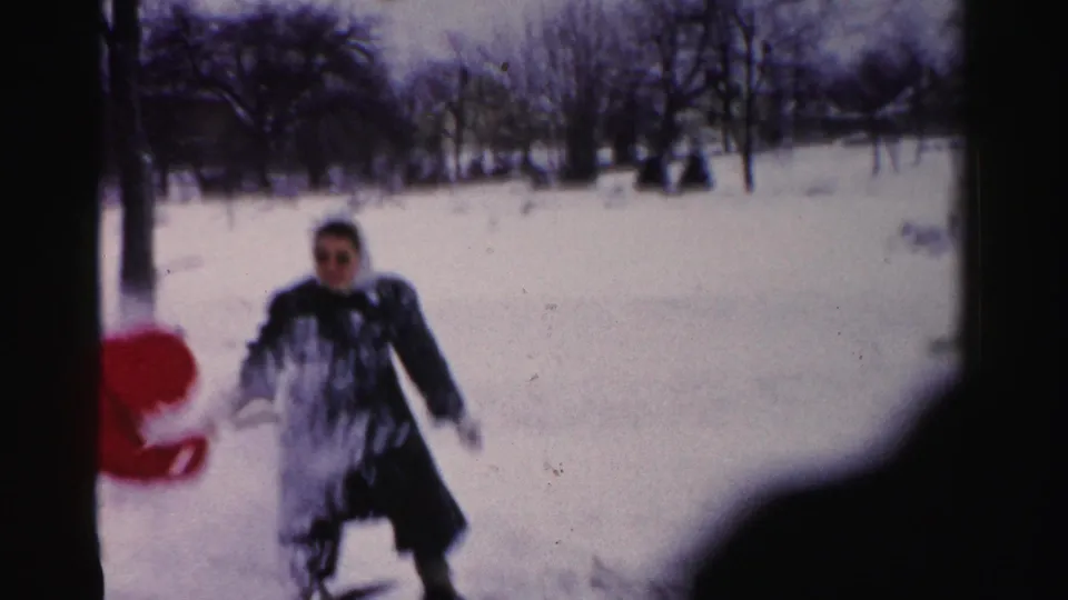 Adult in dark coat facing camera in a snow-covered residential yard, appearing to wear dark-framed glasses; consistent with mid-1950s Albany home movie footage.