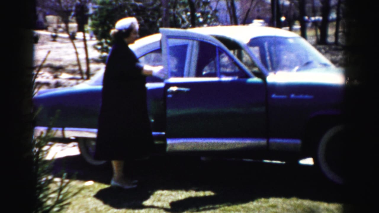 Woman in dark coat and white hat holds car door of a 1950s American sedan in a suburban setting, Albany, New York area, circa 1955–56.