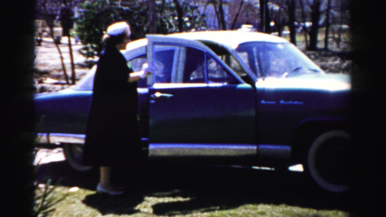 Woman in dark coat and white hat at the open door of a 1950s American sedan; bare deciduous trees and suburban backdrop, Albany NY area, c. 1955–56.