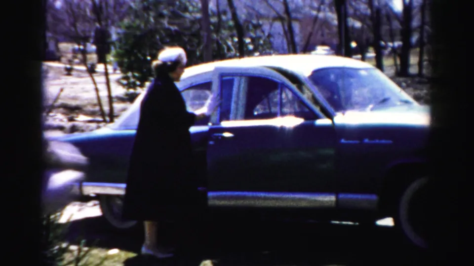 Woman in dark coat at open car door of a 1950s American sedan; bare-branched trees and suburban lawn consistent with late autumn or early spring, Albany NY area, c. 1955–56.