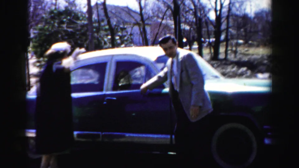 A woman in a dark coat and a man in a light suit meet at the open door of a 1950s American sedan in a suburban setting, Albany, New York area, circa 1955–56.