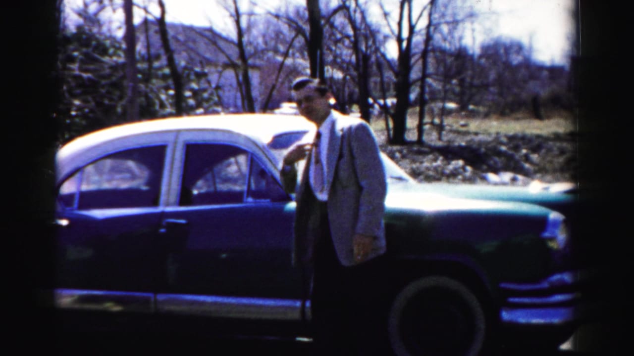 Man in light sport coat beside a large two-tone American sedan with whitewall tires, bare deciduous trees; Albany, New York, mid-1950s 8mm home movie.