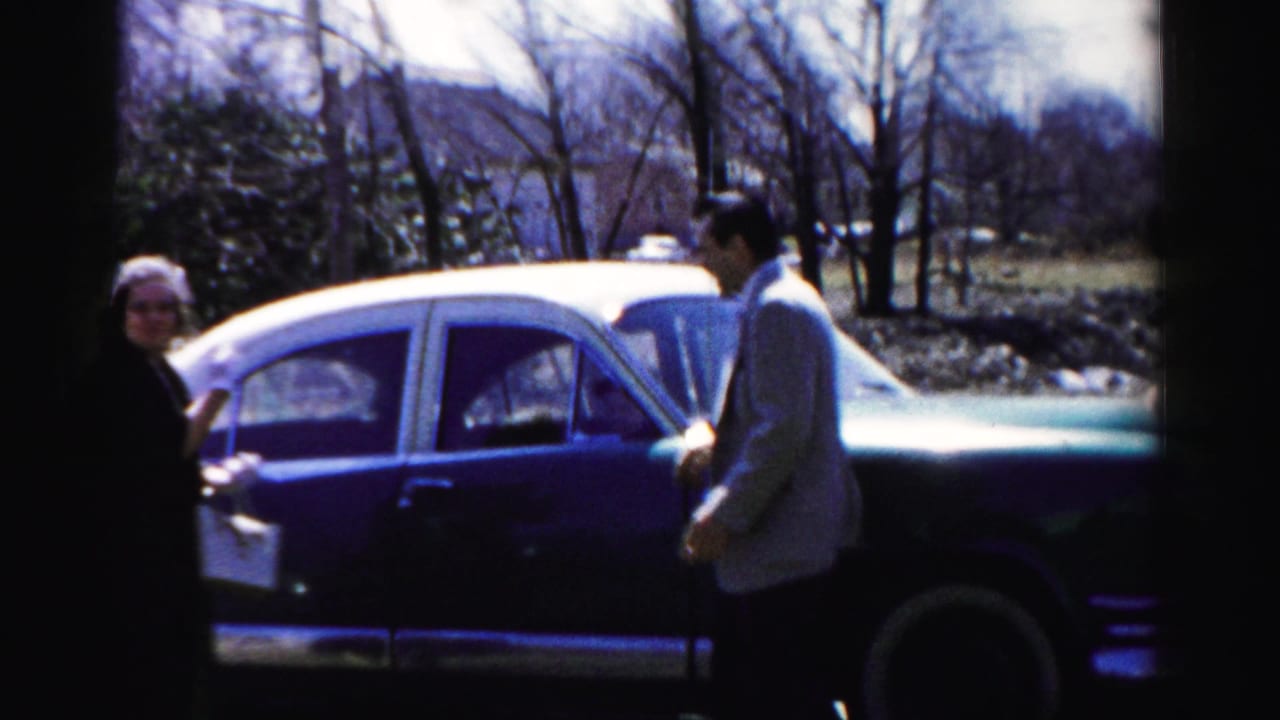 Man in light jacket and woman in dark coat facing each other beside a large two-tone American sedan.