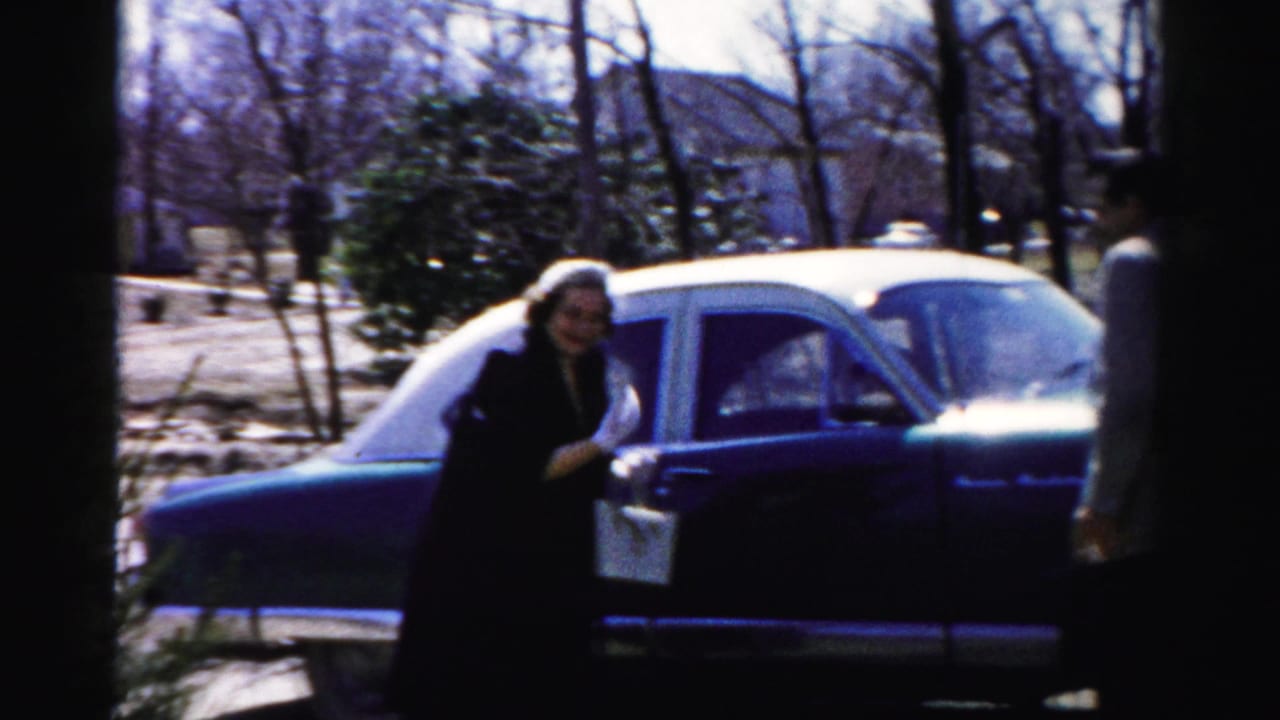 Woman in dark coat near car door in foreground, man partially visible at right; mid-1950s suburban setting consistent with Albany, New York 8mm home movie.