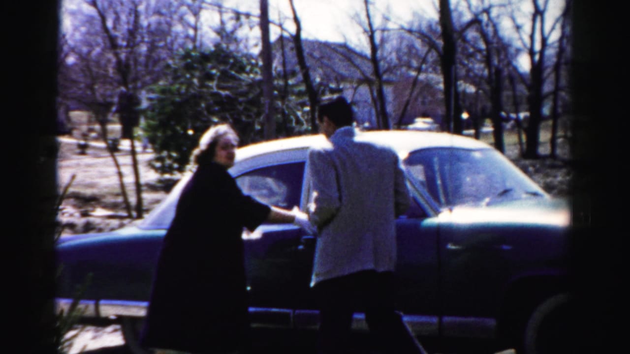 Man in light jacket and woman in dark coat alongside a large American sedan; mid-1950s home movie consistent with Albany, New York metadata.
