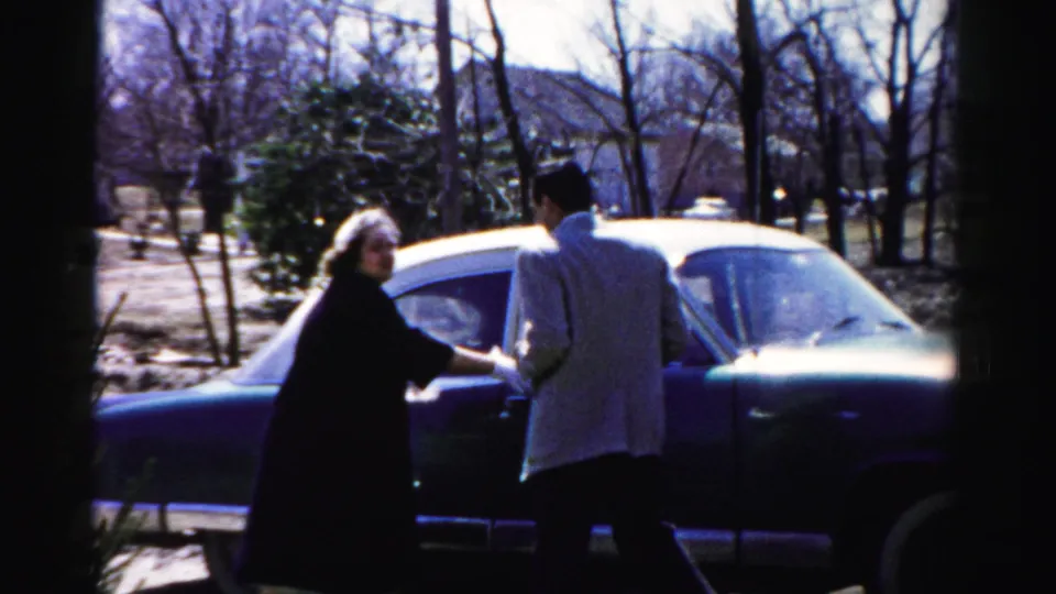 Man in light jacket and woman in dark coat alongside a large American sedan; mid-1950s home movie consistent with Albany, New York metadata.