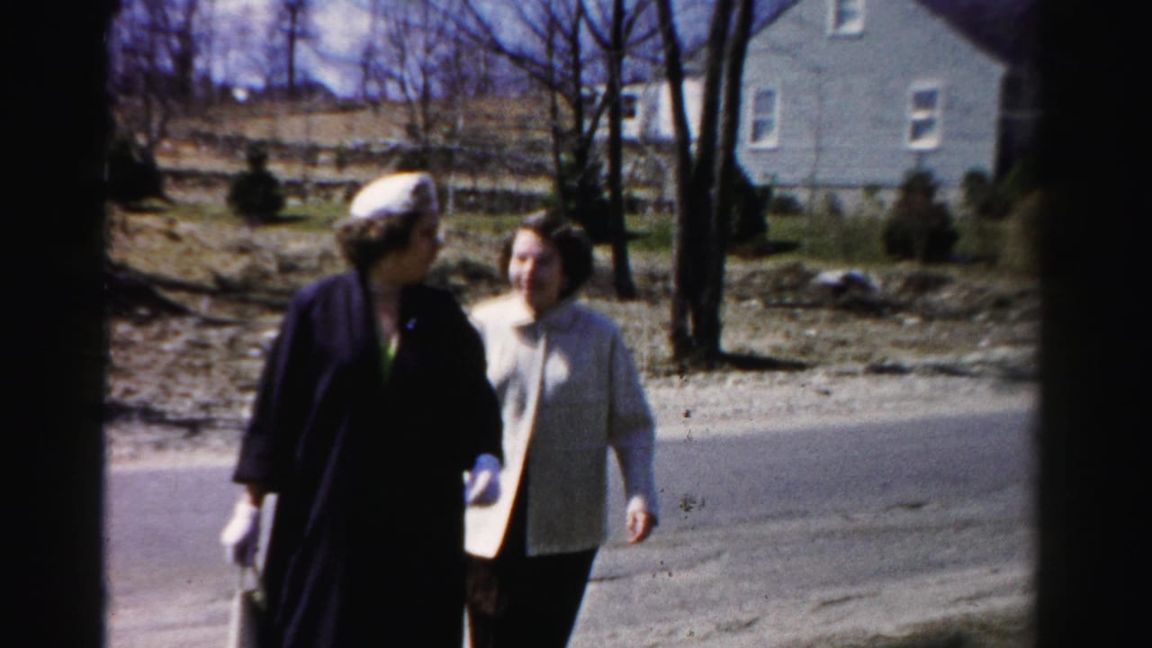 Two women walk and converse on a suburban road; the nearer woman wears a dark coat, beret-style hat, and white gloves.