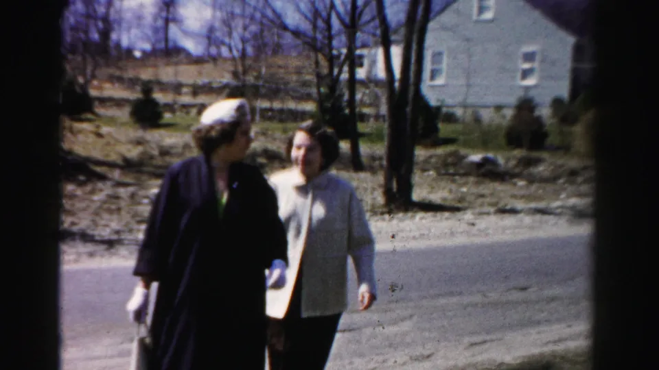 Two women in mid-century dress walk together on a suburban road, one wearing a beret and white gloves, with a residential house and bare trees behind them.