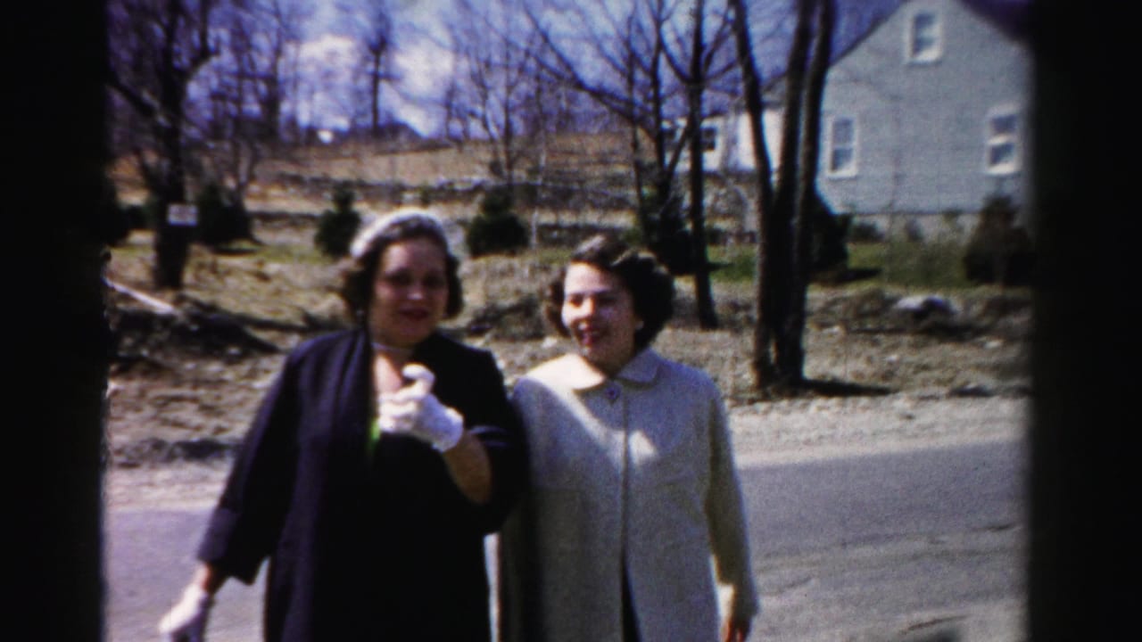 Close view of two women facing the camera and smiling on a suburban road; the woman on the left raises a white-gloved hand toward the lens.
