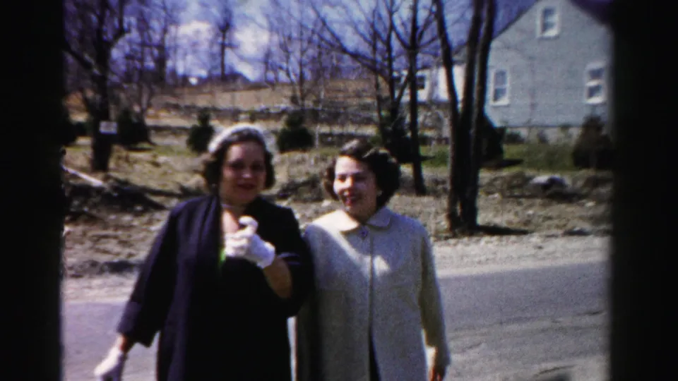Close view of two women facing the camera and smiling on a suburban road; the woman on the left raises a white-gloved hand toward the lens.