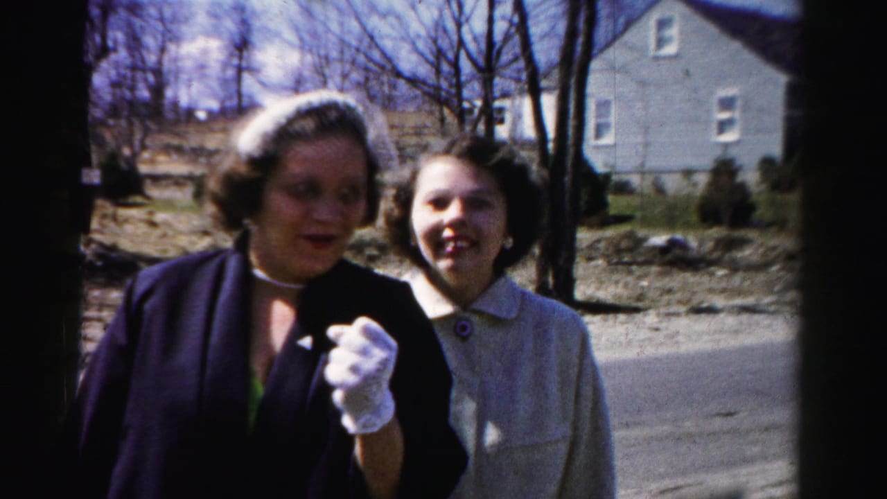 Tight framing of two women laughing and speaking at the camera on a suburban road; one's white-gloved hand is prominent in the lower foreground.