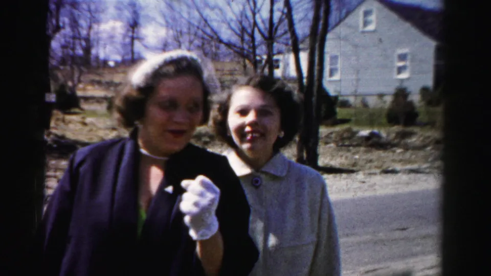 Tight framing of two women laughing and speaking at the camera on a suburban road; one's white-gloved hand is prominent in the lower foreground.