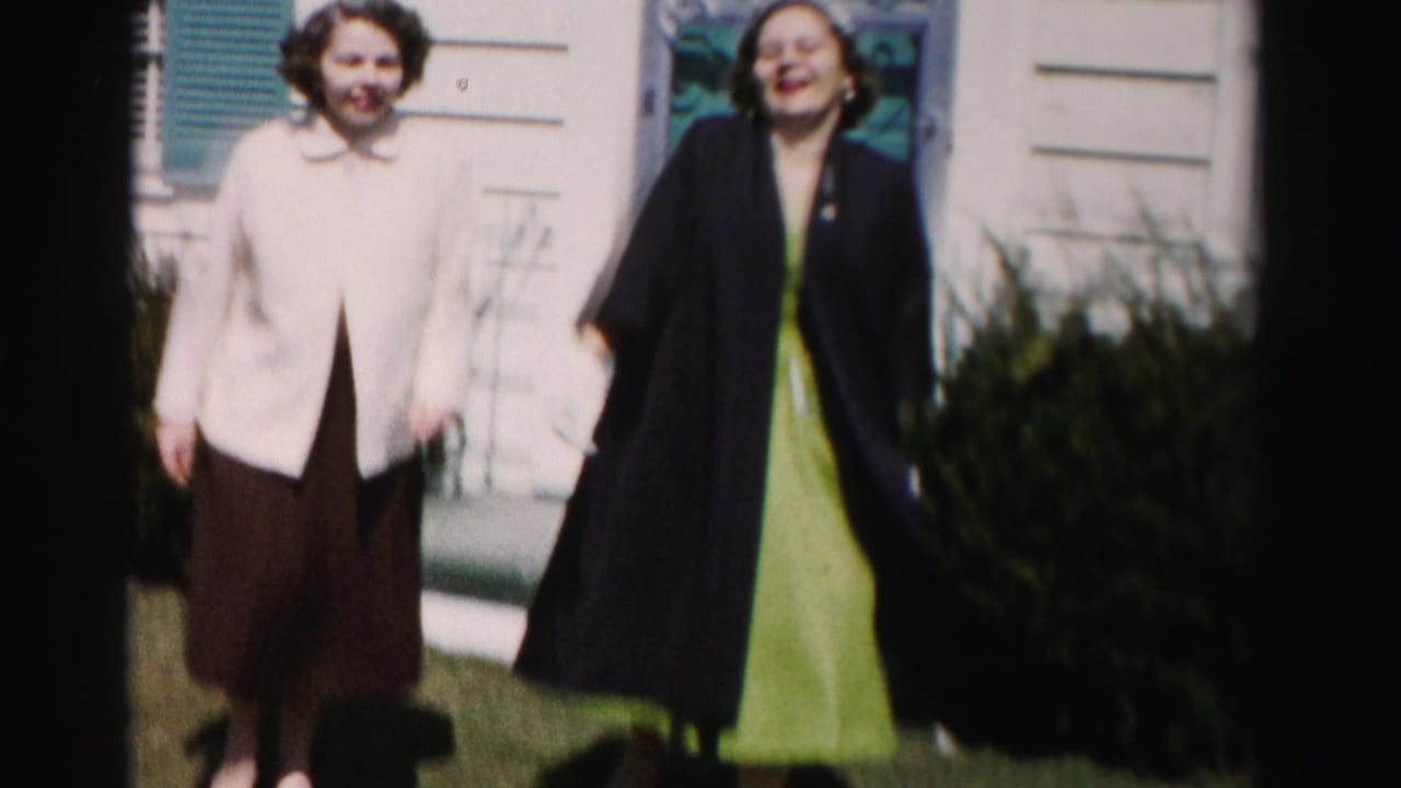 Two smiling women in mid-1950s dress — one in a cream jacket and brown pleated skirt, the other in a dark cape coat over a green dress — stand on a suburban lawn, Albany, New York, circa 1955–1956.