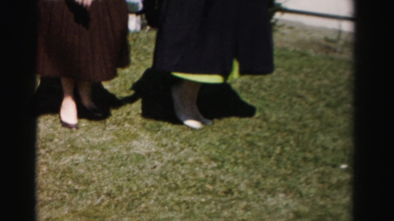 Close shot of two women's lower legs and feet in heeled shoes on a grass lawn; a partial third figure is visible at frame right.