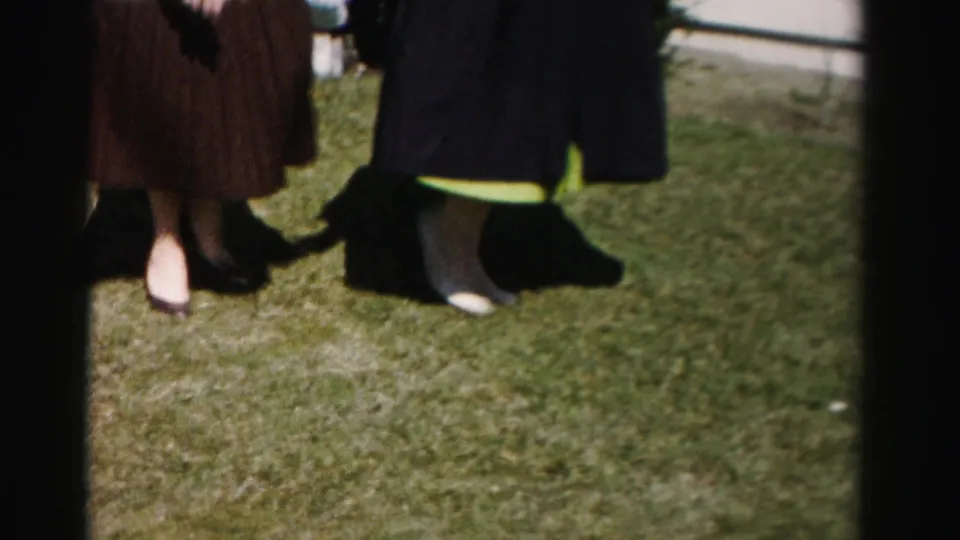 Close shot of two women's lower bodies on a manicured lawn — brown pleated skirt and dark coat with green dress hem visible — with a partial third figure at frame right, circa mid-1950s.