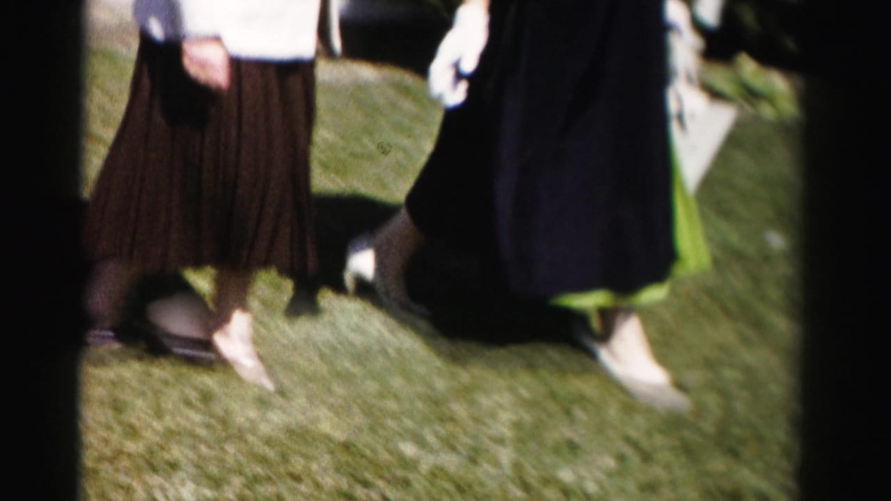 Mid-body close-up of two women in mid-1950s dress on a lawn — brown pleated skirt at left, dark coat with green dress at right — white clutch purse visible in hand.