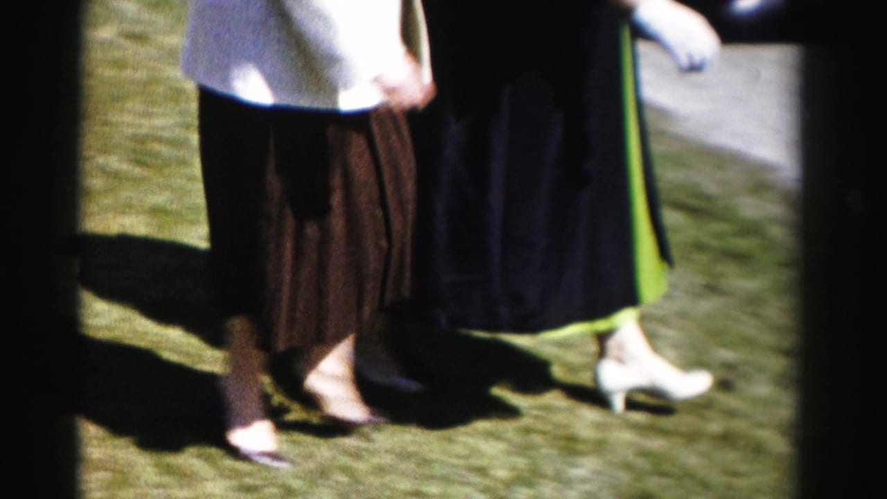 Lower-body shot of two women mid-stride on a lawn — brown pleated skirt on left, dark coat with green dress hem on right — white kitten-heel pumps visible, pavement edge at frame right.