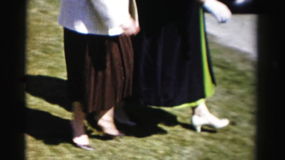 Lower-body shot of two women mid-stride on a lawn — brown pleated skirt on left, dark coat with green dress hem on right — white kitten-heel pumps visible, pavement edge at frame right.