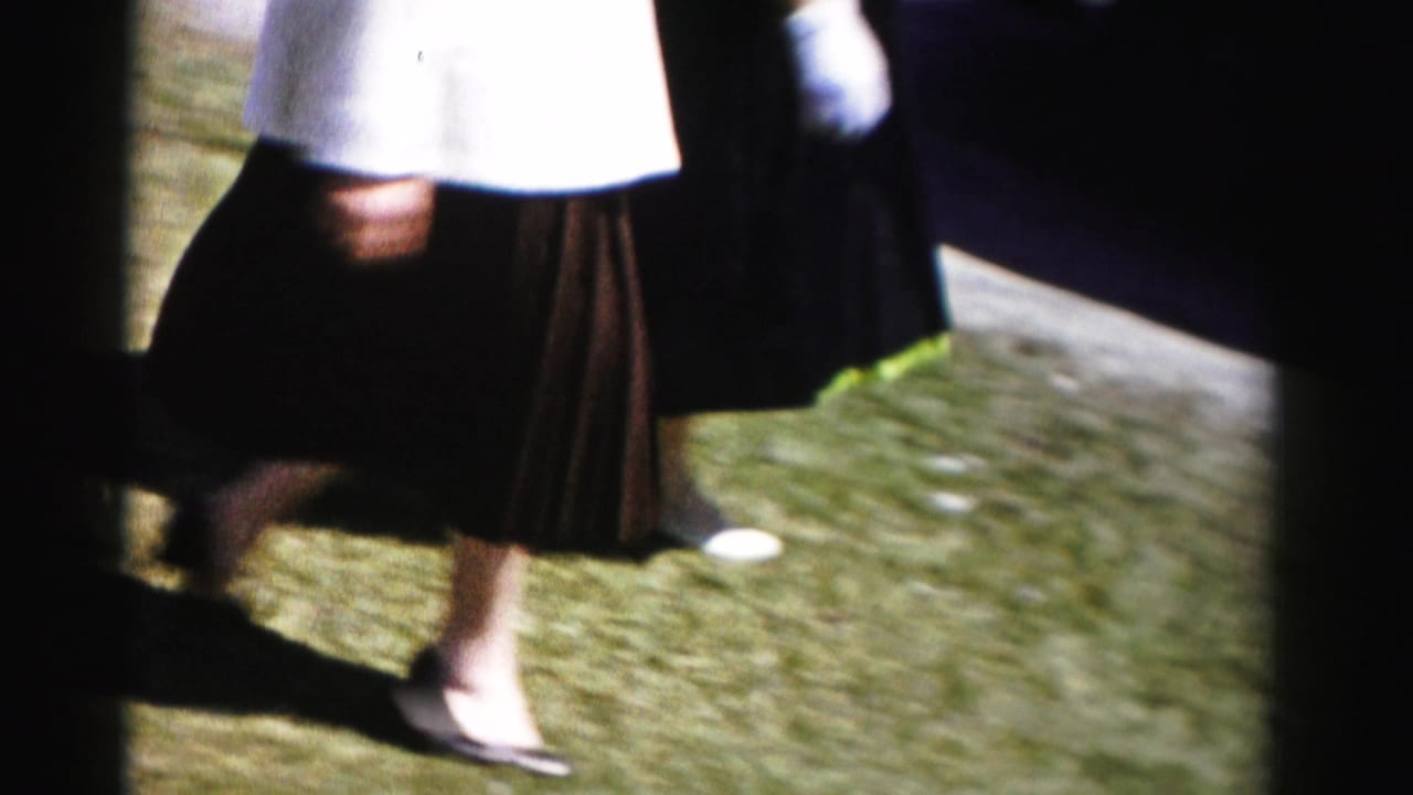 Lower body of a woman in a white top and dark pleated skirt walking across a sunlit green lawn, wearing dark heeled shoes.