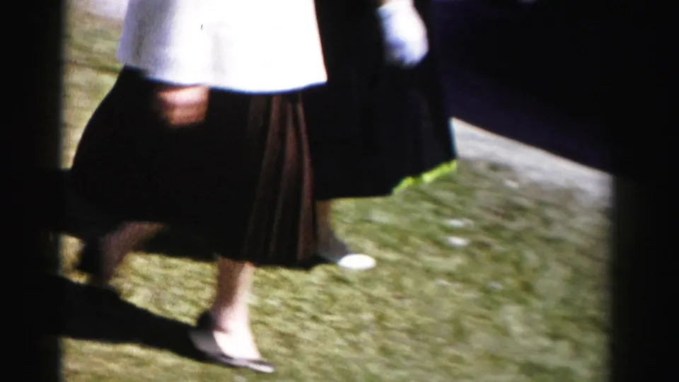 Close-up of a woman's lower body in mid-1950s formal attire — white jacket, dark pleated skirt, dark heeled pumps — crossing a suburban lawn in color 8mm home-movie footage.