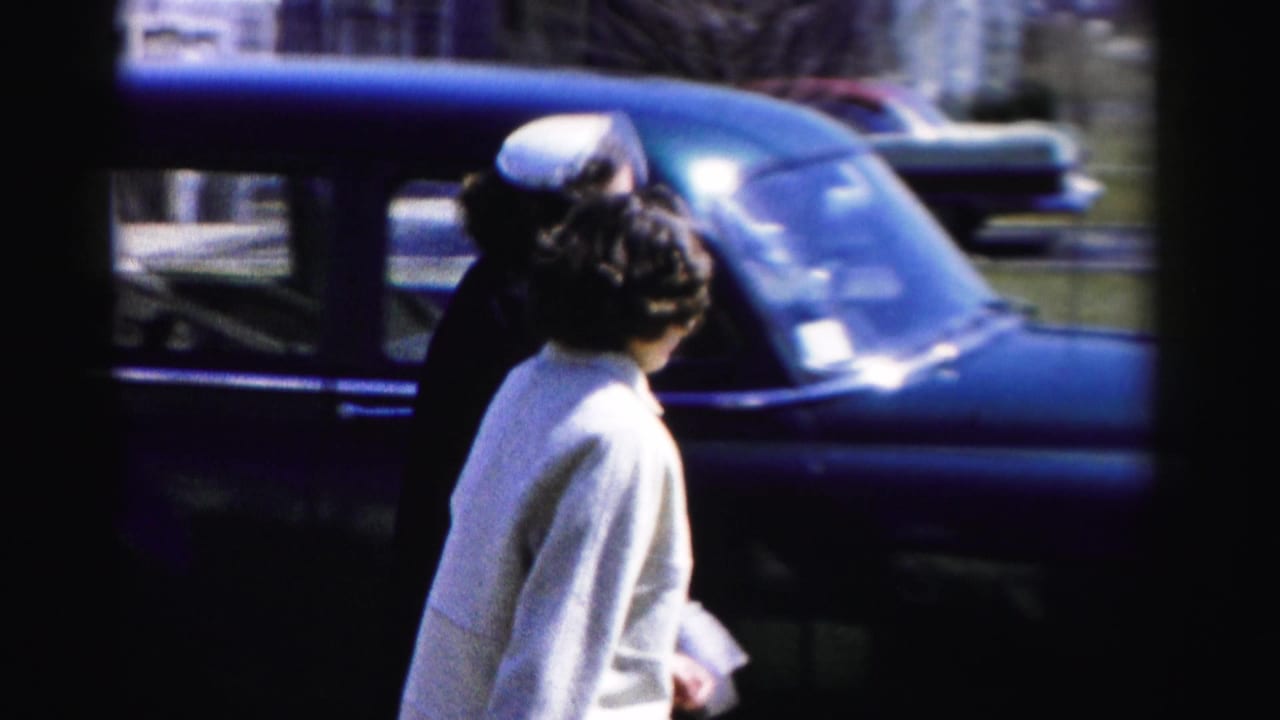 A woman in a white jacket with dark hair pinned up stands near a rounded-body mid-1950s dark-blue automobile, photographed from behind in color 8mm home-movie footage, Albany, NY area.