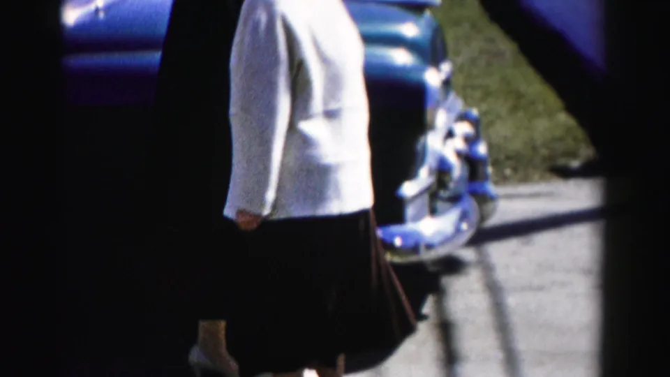 A woman in 1950s formal attire — white jacket, dark pleated skirt — walks past a vintage dark-blue automobile in a suburban setting, with a small figure partially visible near the bumper.