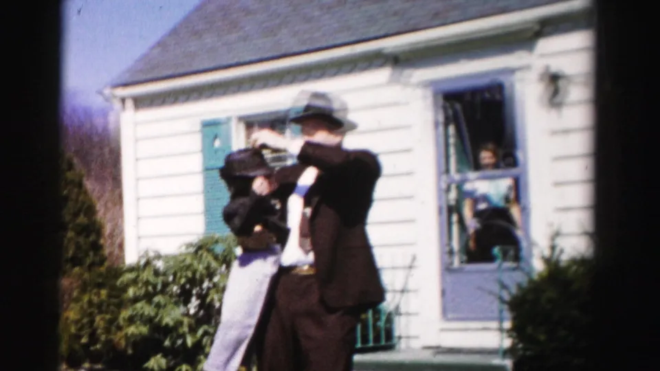 A man in a dark suit and fedora-style hat lifts a young child in front of a white clapboard mid-century house with teal shutters; a second person is visible inside the window, Albany, NY area, mid-1950s.