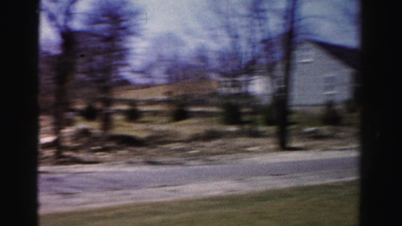 Motion-blurred 8mm pan across a mid-1950s suburban neighborhood with bare deciduous trees, a road, and residential buildings.