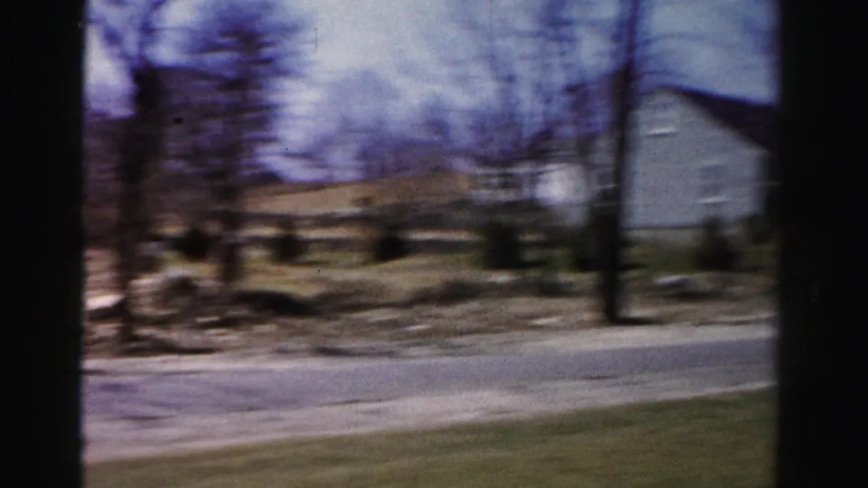 Motion-blurred 8mm pan across a mid-1950s suburban neighborhood with bare deciduous trees, a road, and residential buildings.